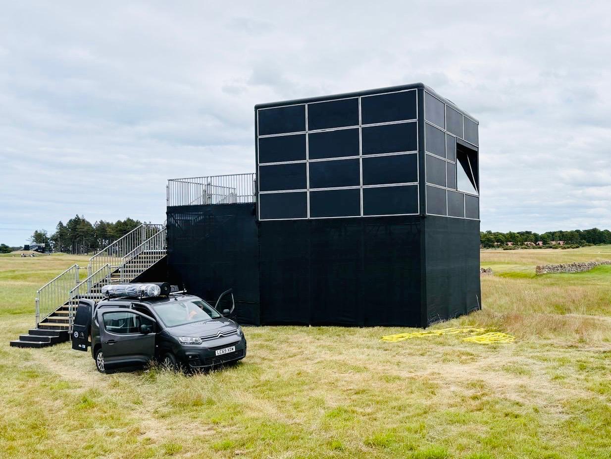 Black-clad scaffold camera tower with access stairs and a van on a UK golf course